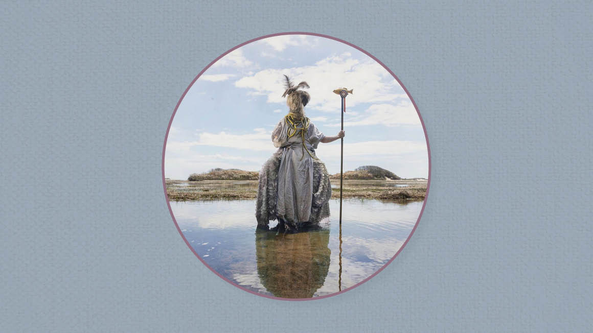 Costumed, masked figure holding a fishing spear stands in shallow water