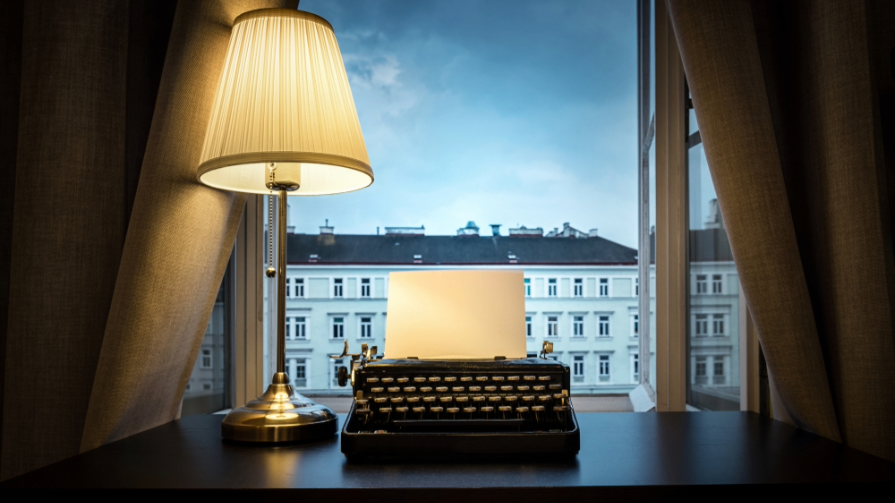 A typewriter sits on a desk in front of a window.