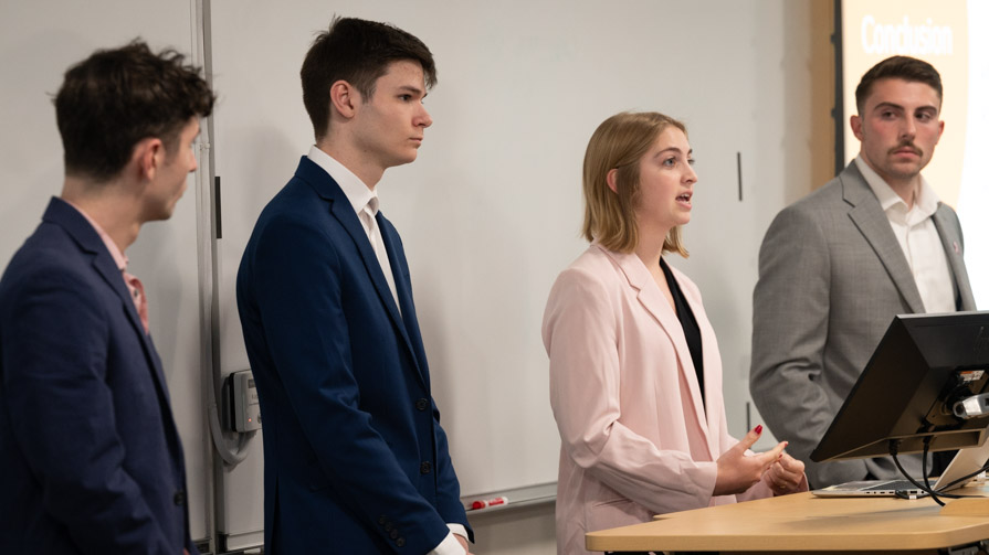 Four students stand in the front of a Bryant classroom and present their work.