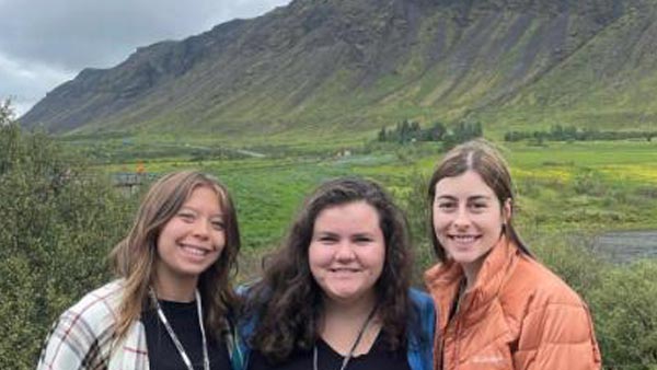 3 female students pose for a photo in front lush green mountains in Iceland.